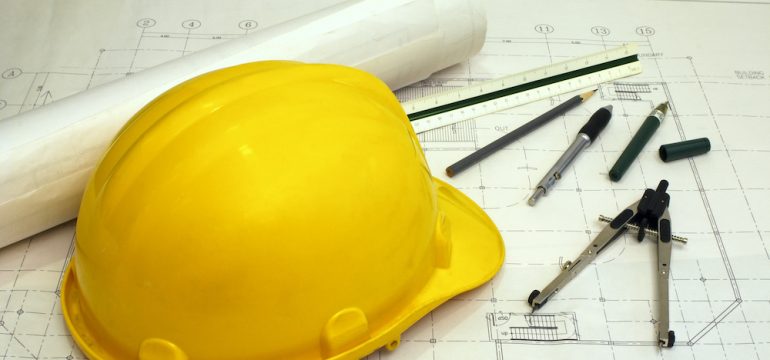 Structural engineer equipment including a yellow hard hat and tools laying on a blueprint of a house.