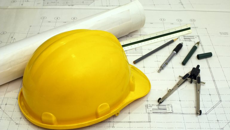 Structural engineer equipment including a yellow hard hat and tools laying on a blueprint of a house.