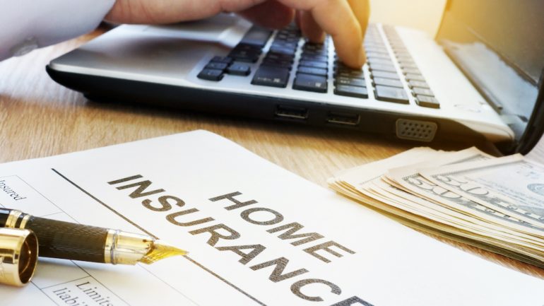 Homeowners insurance form on a table with an agent's hands typing on a laptop computer. A stack of money is also on the desk to represent money saved on homeowners insurance.