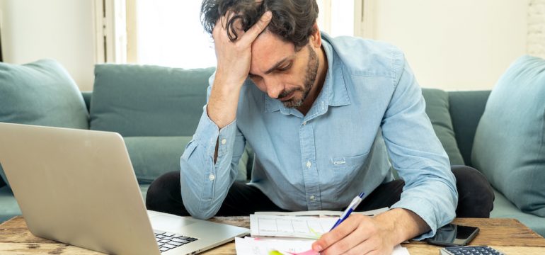 Young man worried about financial relief during Covid-19 pandemic. Working with laptop at home looking at bills.