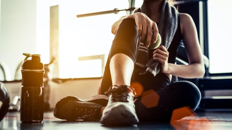 Woman sitting and resting after fitness training in gym with protein shake or drinking water on floor. Relax after training concept.