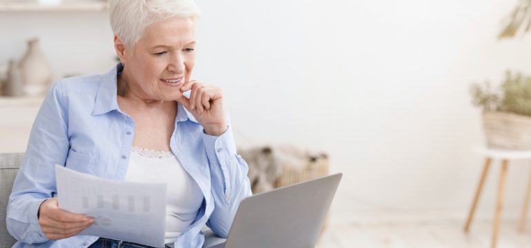 Smiling retired woman holding asset-based mortgage documents. Using a laptop at home, sitting on couch In living room.