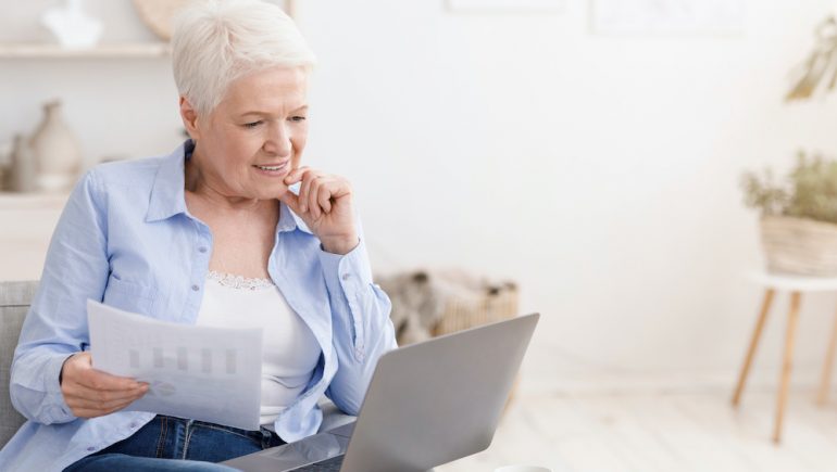 Smiling retired woman holding asset-based mortgage documents. Using a laptop at home, sitting on couch In living room.