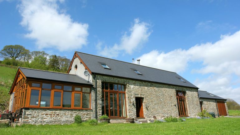 Newly converted residential barndominium with a slate roof in rural countryside against a blue sky with clouds.