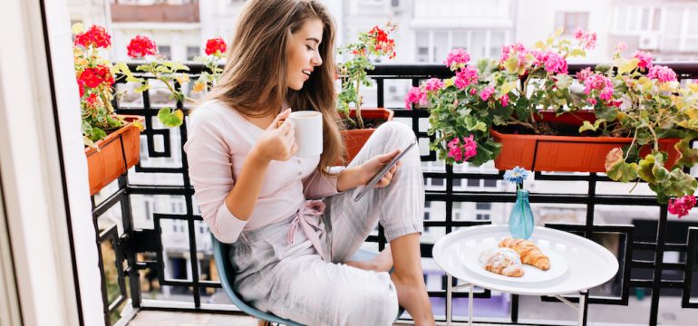 Attractive girl with long hair in having breakfast on balcony in the morning in city. She holds a cup, reading on tablet.