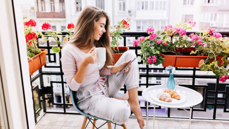 Attractive girl with long hair in having breakfast on balcony in the morning in city. She holds a cup, reading on tablet.