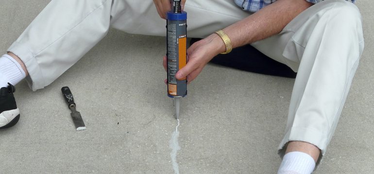 Man filling a cracked concrete in patio block with a caulking gun and putty knife.