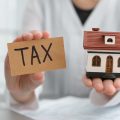 Woman holding house model and card with word TAX. Presents the concept of a tax lien on a house.