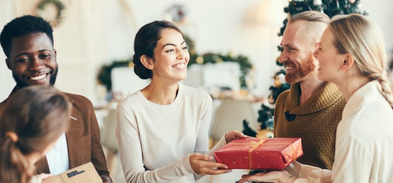 Group of elegant young people exchanging gifts with overnight guests and smiling cheerfully during holiday party.