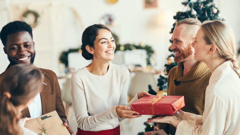 Group of elegant young people exchanging gifts with overnight guests and smiling cheerfully during holiday party.