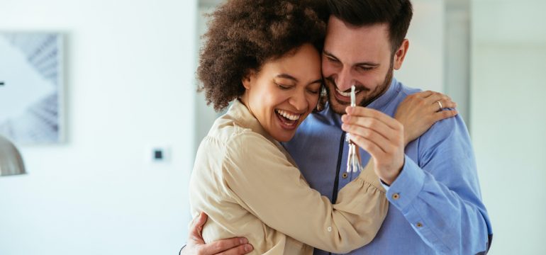 Young home buyers in their 20s showing key with their arms around each other.