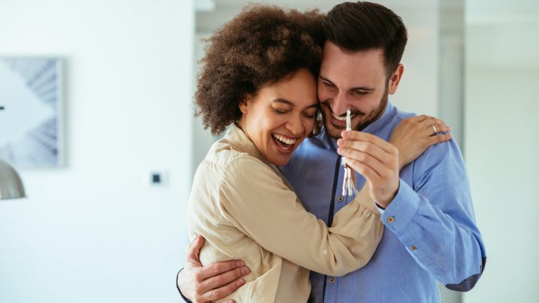 Young home buyers in their 20s showing key with their arms around each other.