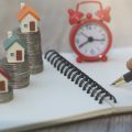 Planning to buy a home for the short term. Calculating in a notebook if it makes sense with a clock and house models sitting on a stack of coins to demonstrate concept.