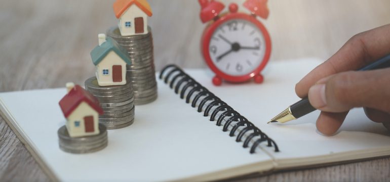 Planning to buy a home for the short term. Calculating in a notebook if it makes sense with a clock and house models sitting on a stack of coins to demonstrate concept.