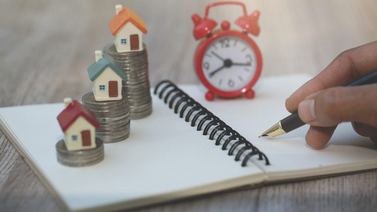 Planning to buy a home for the short term. Calculating in a notebook if it makes sense with a clock and house models sitting on a stack of coins to demonstrate concept.