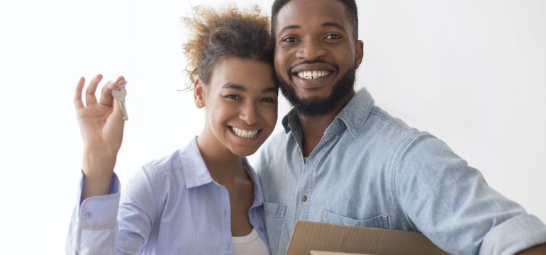 Young woman showing a house key with her significant other holding moving box in their new living room.