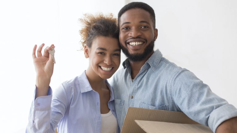 Young woman showing a house key with her significant other holding moving box in their new living room.