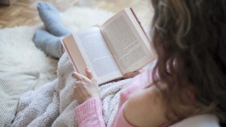 Coziness at home with the Hygge lifestyle. Photo of girl doing reading a book covered with a warm blanket.