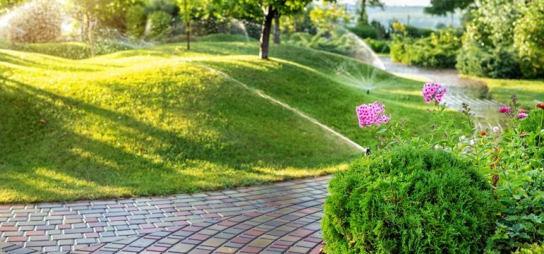 Home irrigation system with different sprinklers installed under the lawn. Landscape design with grassy hills and fruit trees irrigated with smart automatic sprinklers at sunset.