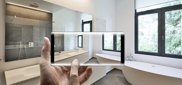 Cell phone in a man's hands taking a video home tour in a staged bathroom with windows looking out towards a garden.