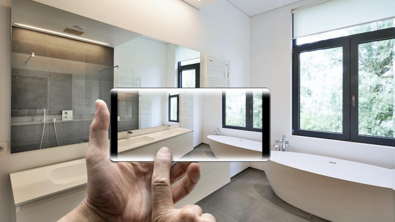 Cell phone in a man's hands taking a video home tour in a staged bathroom with windows looking out towards a garden.