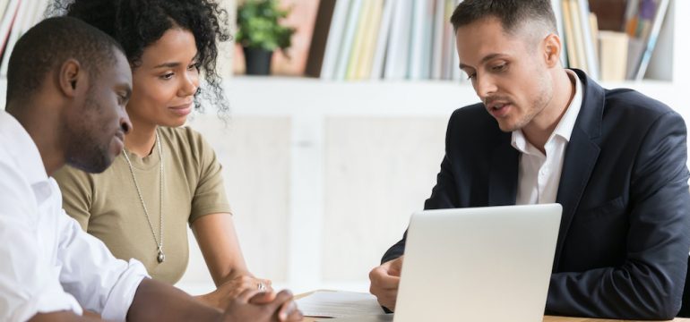 Male real estate agent meeting with millennial couple showing properties on laptop. The agent can only answer questions allowed within Fair Housing Laws.