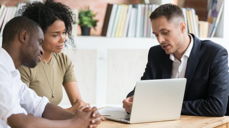 Male real estate agent meeting with millennial couple showing properties on laptop. The agent can only answer questions allowed within Fair Housing Laws.