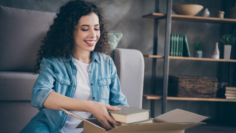 Attractive, cheerful wavy-haired female unpacking books as she moves into a modern loft. Shows trend of single women buying their own homes.