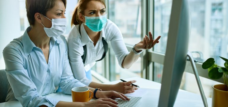Female colleagues wearing face masks while working on a computer and communicating about possible coronavirus cyber scams via an e-mail.