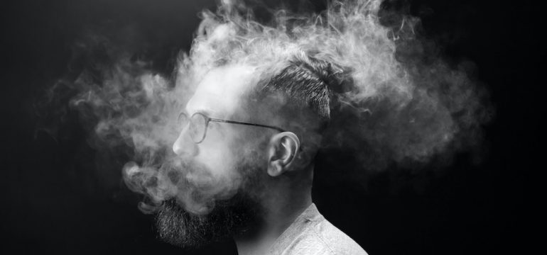 Portrait of a bearded, stylish man with cigarette smoke enveloping his head. Concept of secondhand smoke.