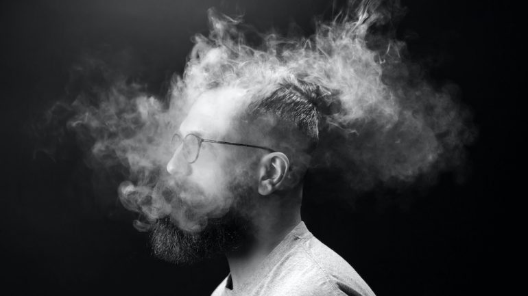 Portrait of a bearded, stylish man with cigarette smoke enveloping his head. Concept of secondhand smoke.