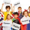 Expat children wrapped in flags of USA and European nations, holding greeting signs in different foreign languages.