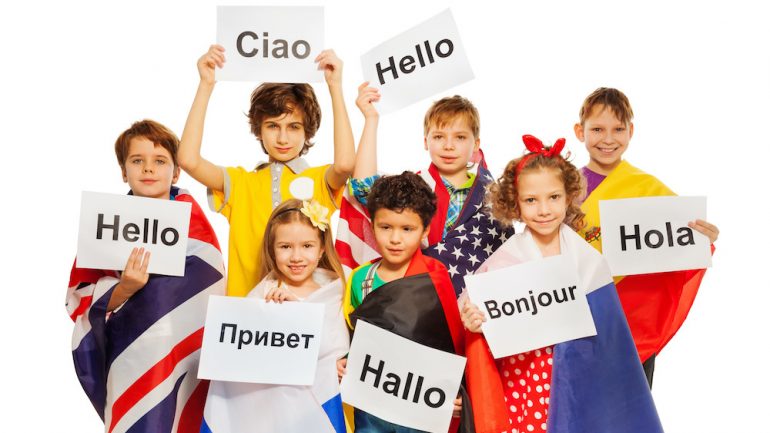 Expat children wrapped in flags of USA and European nations, holding greeting signs in different foreign languages.