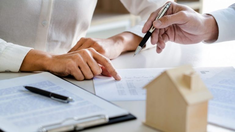 Real estate agent reviewing a mortgage loan contingency with a home buyer customer. Showing discussion over contract on a table with a wooden model home.