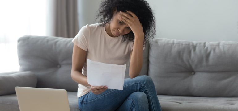 Stressed young woman holding papers sitting on a sofa wondering how to avoid foreclosure on her home.