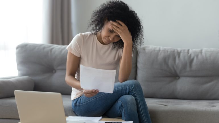 Stressed young woman holding papers sitting on a sofa wondering how to avoid foreclosure on her home.