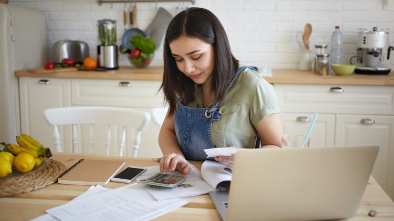 Indoor shot of attractive young female doing calculations, managing monthly bills, sitting at kitchen table and smiling.