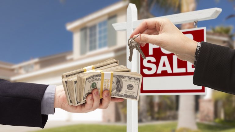 Real estate agent handing over the keys as home buyer is handing over all-Cash for home purchase. House with "For Sale Sign" in background.