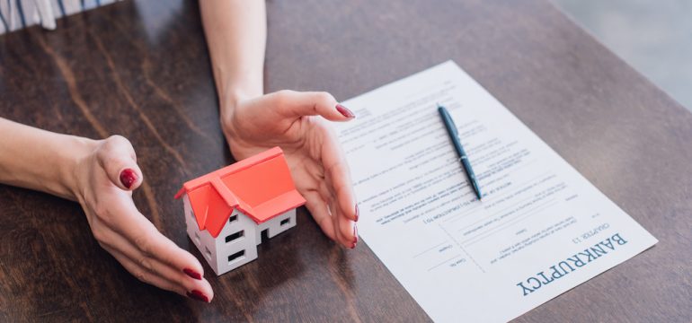 Cropped view of female hands near house model, paper with bankruptcy lettering and pen on table. Homeownership after bankruptcy concept.