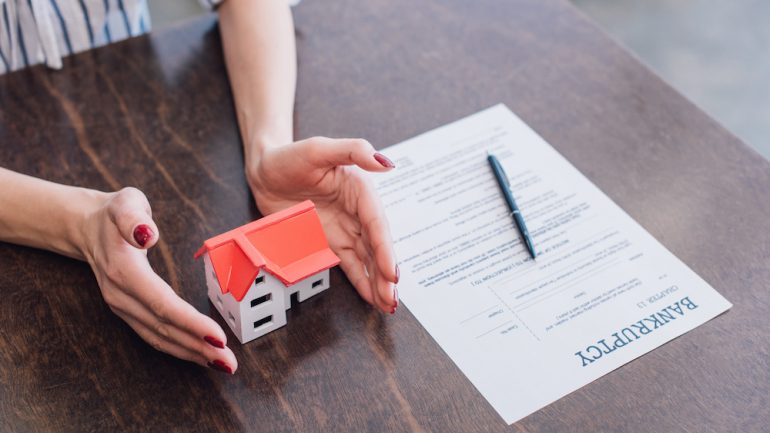 Cropped view of female hands near house model, paper with bankruptcy lettering and pen on table. Homeownership after bankruptcy concept.