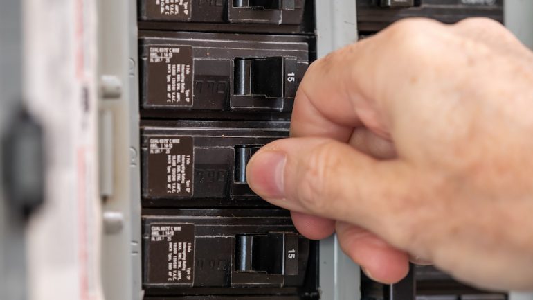 Male electrician turning off power for electrical outlet at circuit breaker box. Resetting tripped breaker in residential electricity power panel.