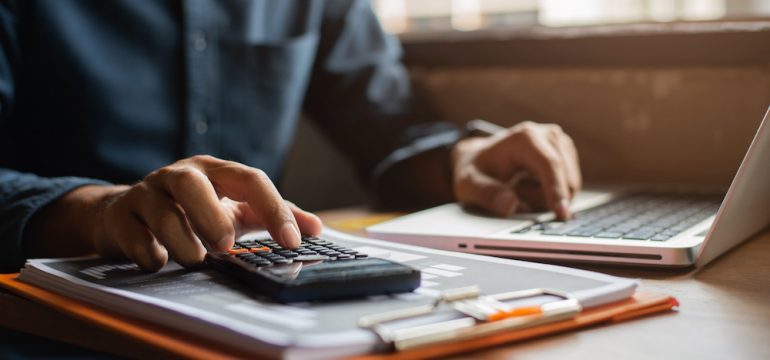 Gentleman's hand calculating the closing costs before selling his home at desk in an office.