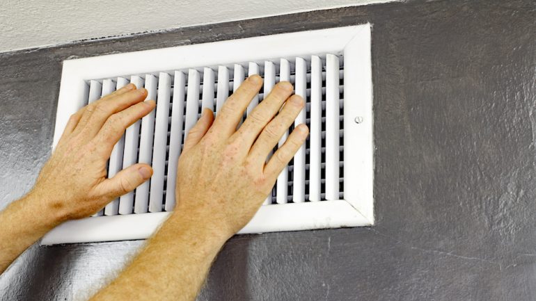 A pair of adult male hands feeling the flow of air coming out of an air vent on a wall near a ceiling before shutting vent to save on energy.