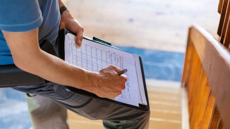A close up and high angle view of a professional male wearing blue t-shirt, writing out forms during a FHA home inspection, standing on stairs with copy-space.