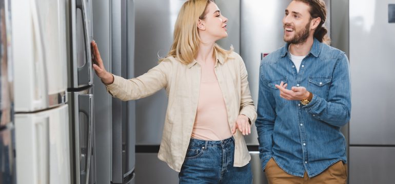 Smiling boyfriend pointing with hand and girlfriend touching appliance finishes on refrigerators in a home appliance store.