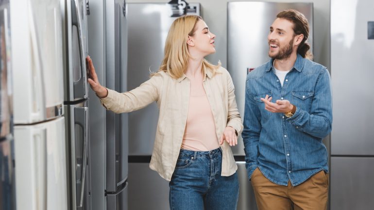 Smiling boyfriend pointing with hand and girlfriend touching appliance finishes on refrigerators in a home appliance store.