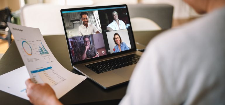 Woman holding a business report showing work from home office with virtual meeting technology on laptop computer.