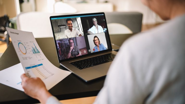 Woman holding a business report showing work from home office with virtual meeting technology on laptop computer.