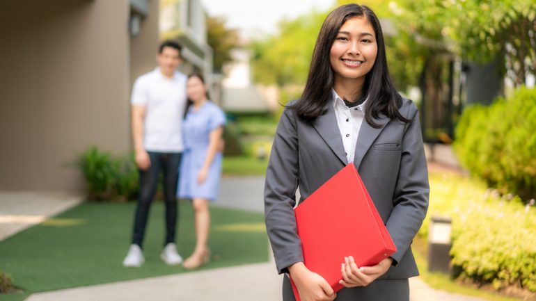 Female real estate agent in a suit holding red file and smiling with a young couple buying a home in front of a house.