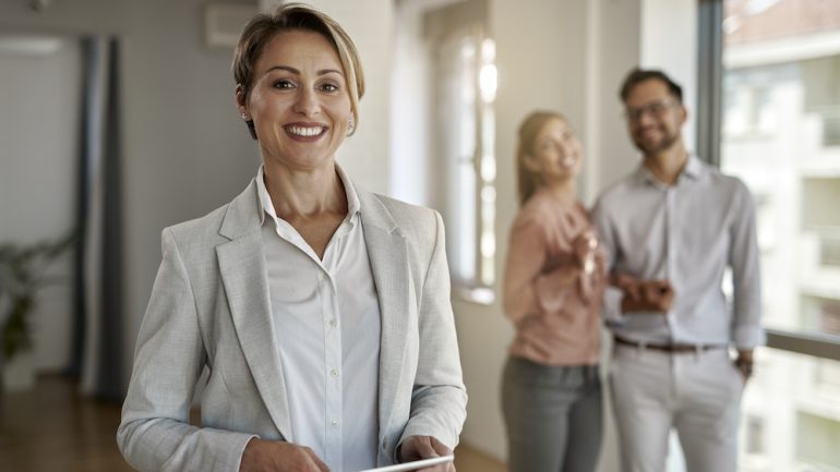 Photo of discount broker real estate agent looking at the camera while her clients are standing in the background.